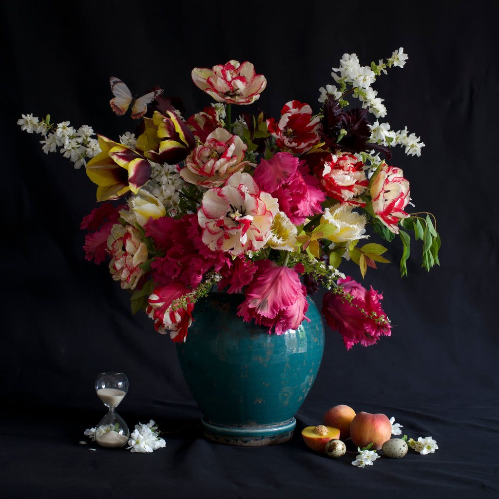 A large bouquet of colourful flowers in the style of still life in a blue ceramic vase including spring blossom and tulips, with fruit, bird eggs, and an hourglass on a black background.


