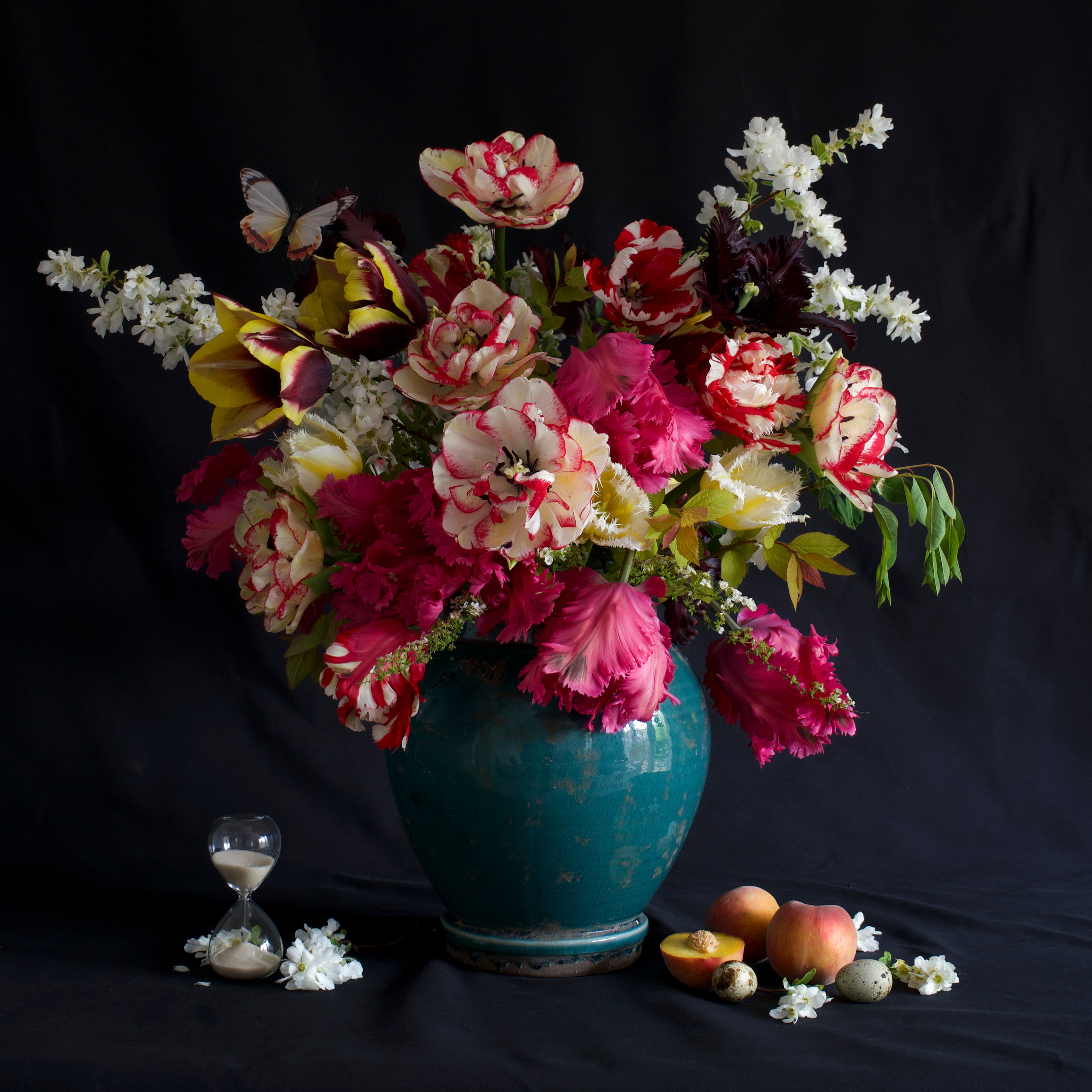 A large bouquet of colourful flowers in the style of still life in a blue ceramic vase including spring blossom and tulips, with fruit, bird eggs, and an hourglass on a black background.


