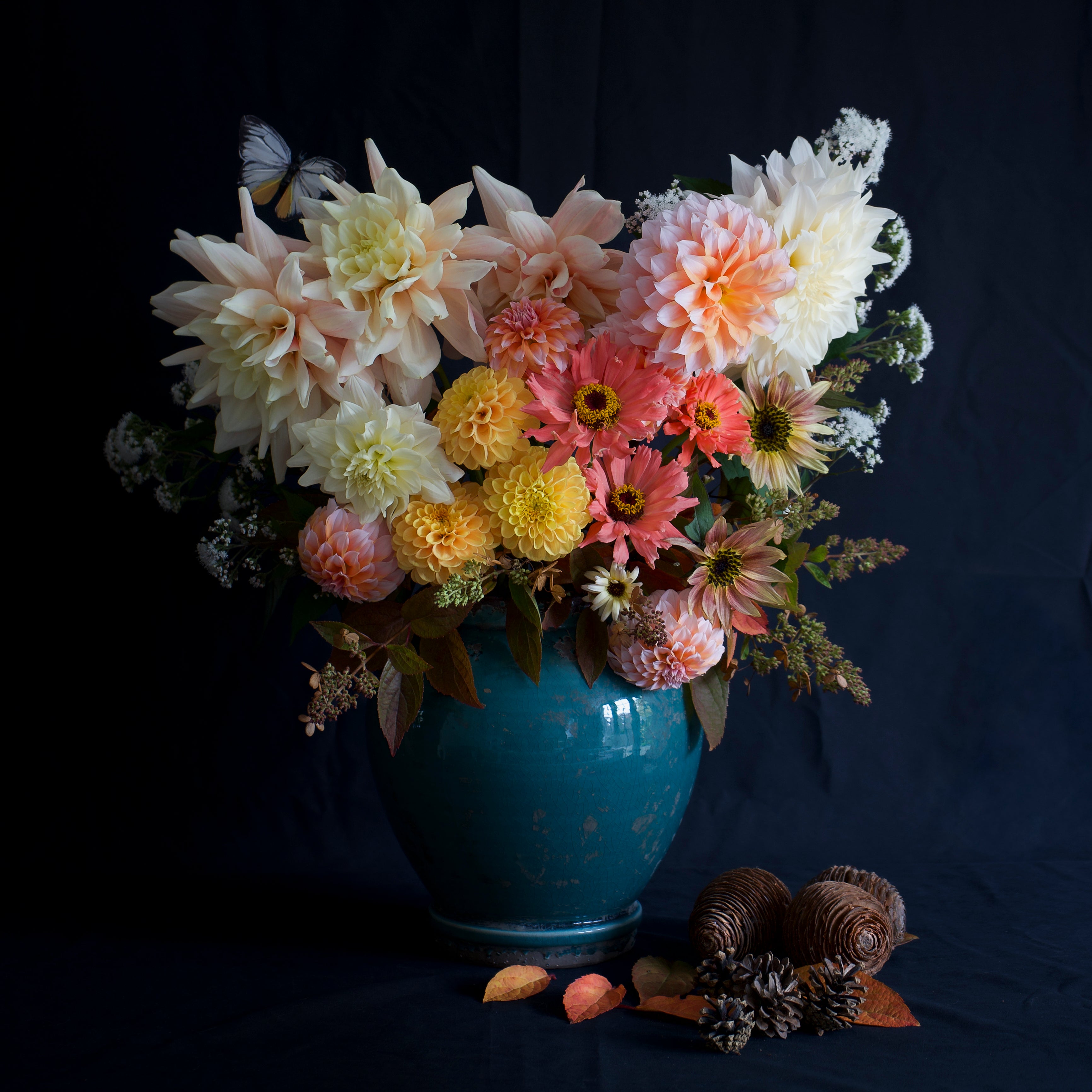A blue ceramic vase filled with a colourful arrangement of large dahlias, zinnias, and sunflowers, with a butterfly perched on a flower, set against a dark background, surrounded by pinecones and fall leaves.