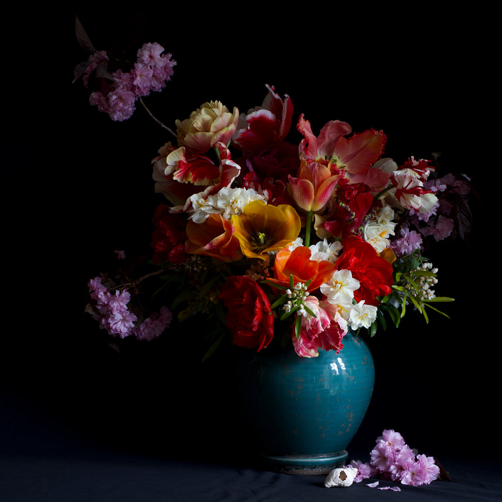 A vibrant bouquet of various colourful flowers in the style of still life including tulips, cherry blossom and narcissus in a blue ceramic vase, with some flowers and petals scattered at the base, against a dark background.